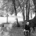 Photographie en noir et blanc de Gilles Colliard assis en forêt prêt d'une croix ancienne, entourée d'une lumière naturelle et apaisante