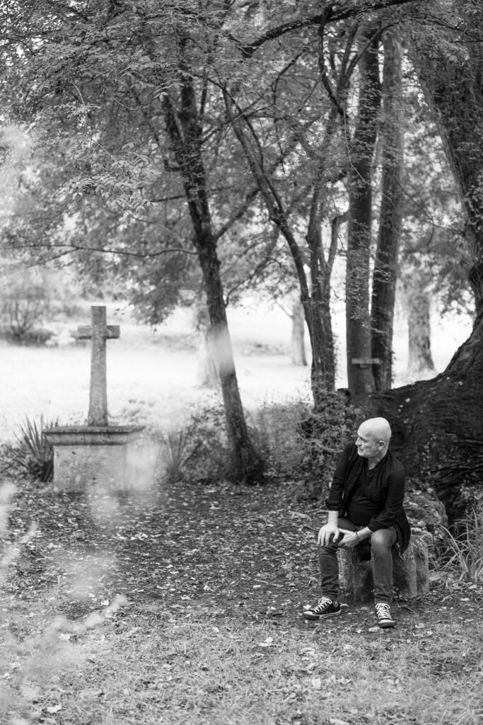 Photographie en noir et blanc de Gilles Colliard assis en forêt prêt d'une croix ancienne, entourée d'une lumière naturelle et apaisante